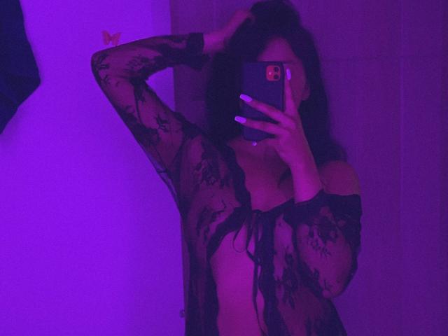 ElizabethMacy with dark hair, wearing a black lace sheer top, exposing her tits, taking a mirror selfie with purple lighting, hand in hair, phone covering her face.