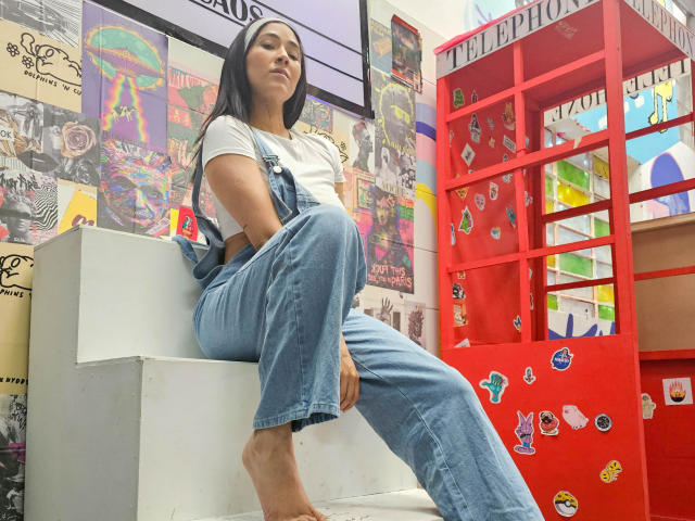 NicoColeman with long black hair, white tee, and blue denim overalls, sitting barefoot on white steps, red phone booth with stickers in background, looking confident.