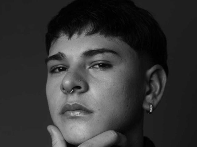 Black-and-white photo of LukeHowlandy with short dark hair, nose ring, and small hoop earring, holding chin, looking slightly to the side with a calm expression.