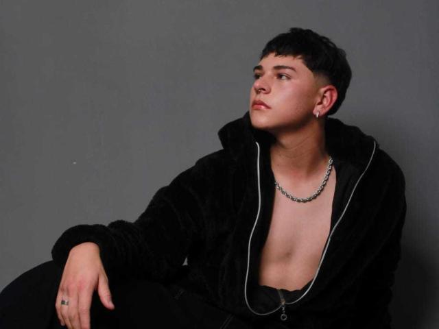 Luke Howlandy, avec ses cheveux noirs courts, portant une veste noire et duveteuse, sans chemise, une chaîne noire, regardant sur le côté, a un petit tatouage sur sa main droite..
