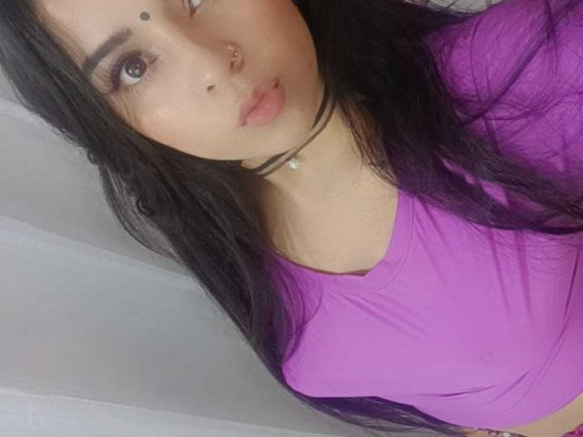 UmaRany with long black hair, purple top, nose ring, and makeup, showing her face and upper body, with a small beauty mark above her right eyebrow. Boobs visible.