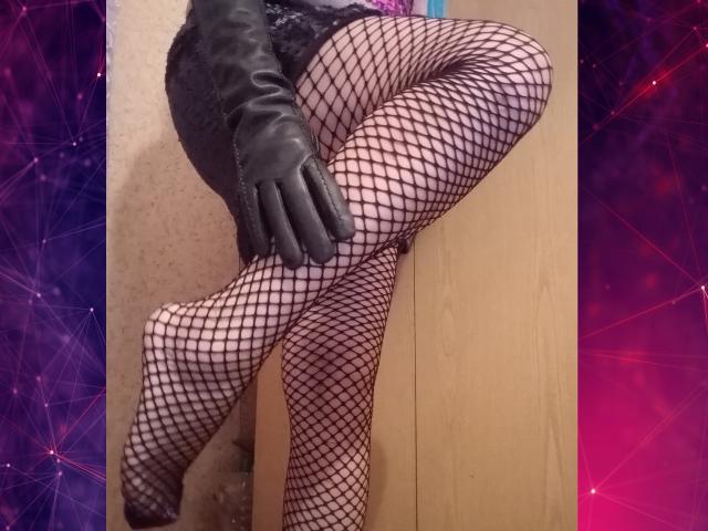 AlinaMistress has her legs crossed, wearing black fishnet stockings and black leather gloves, with a wooden surface behind her. Her feet are pointed, showing off her sexy legs.