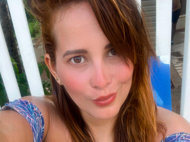 SaritaVelez has light brown eyes, reddish-brown hair, and is wearing a blue patterned top with her tits slightly visible, looking at the camera with a small smile.