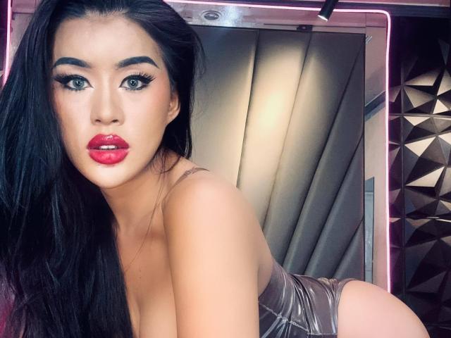QuiinMolly with long black hair, red lipstick, and thick eyeliner, looking at the camera, showing her tits and ass in a shiny, tight, gray top.