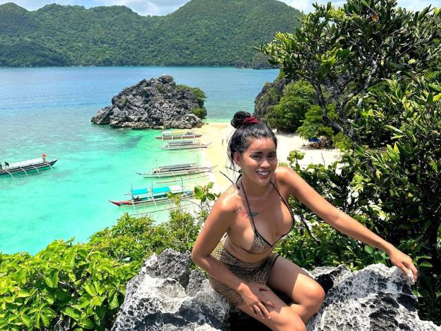 QuiinMolly with small tits and a tan, bare-ass bikini, sitting on rocks, smiling, black hair in a bun, tropical beach with boats, green hills, and clear blue water.