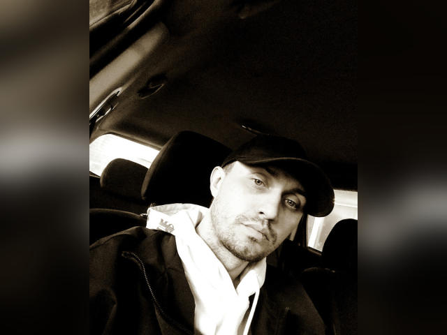 Sepia-toned selfie of Svit with a slight smirk, wearing a dark baseball cap and hoodie, sitting in a car. Svit's face is bearded, looking to the side.