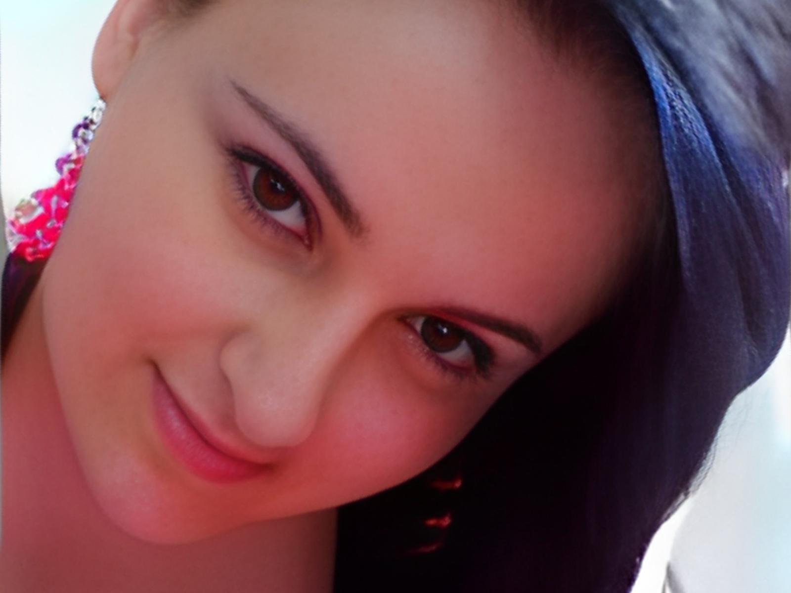 HayleyCam has a cute, slightly flushed face with brown eyes, dark eyebrows, and a small smile. She's wearing a black headscarf with pink, curly-patterned clothes visible.