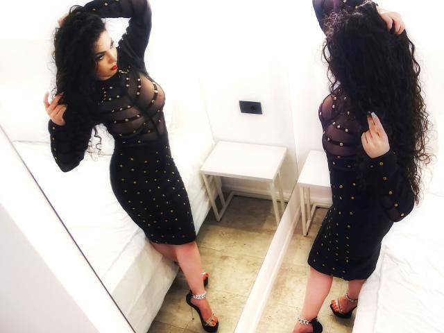 Lynxy with curly black hair, wearing a sheer black dress with gold studs, showing her tits and ass, standing in front of a mirror, in black high heels.