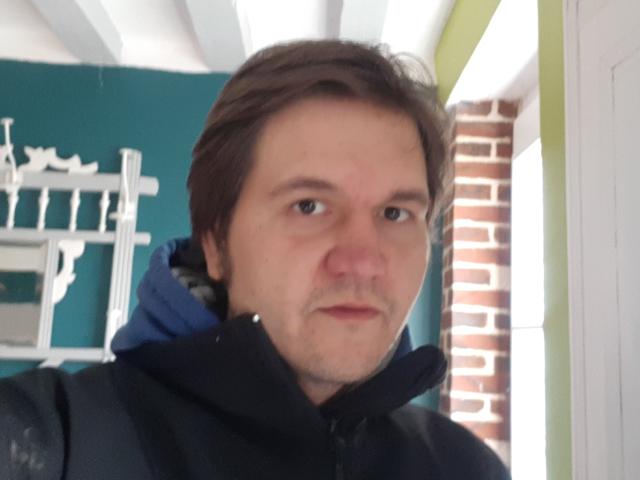 NectarineFad with a slightly flushed face, brown hair, and a black jacket, standing in a doorway with a blue hood visible. Nervous expression, slightly parted lips.