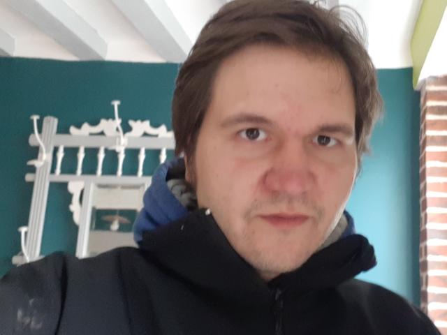 NectarineFad with brown hair, light skin, and a slight mustache, wearing a black jacket, looking slightly annoyed. His blue hoodie is visible behind his jacket.