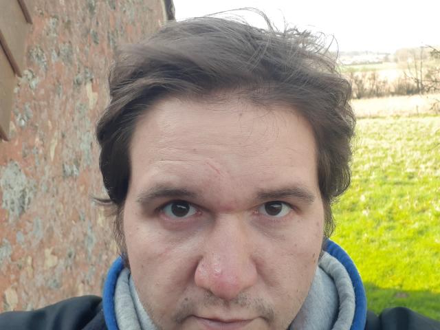NectarineFad tiene un rostro ligeramente desaliñado, con ojos marrones, cabello castaño claro y una pequeña cicatriz en la nariz. Lleva una sudadera azul con cuello gris..