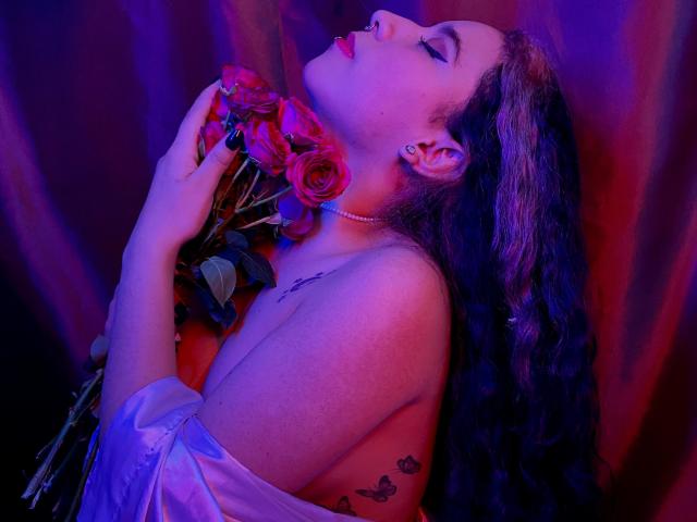 SophieDreamLove met paars en roze neonverlichting, houdt rozen voor haar neus, ogen gesloten, lippenstift op, lang krullend bruin haar, parelketting, jurk met open schouder, vlindertatoe op haar arm..