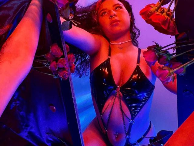 SophieDreamLove with curly brown hair, wearing a shiny black latex bikini, chains across her belly, rose petals around, and a nose ring, looking up with a pouty expression.