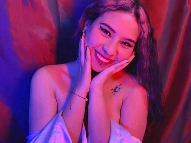 SophieDreamLove has pink and blue lit skin, wavy hair, and a smile showing red lipstick. She's touching her face, showing a crescent moon tattoo on her chest. Boobs.