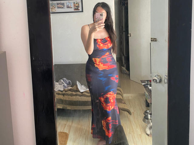 ClaudiaBuenaVista in a tight, strapless, dark blue dress with red and orange flames, standing in front of a mirror, holding a phone, covering her tits with her hand.