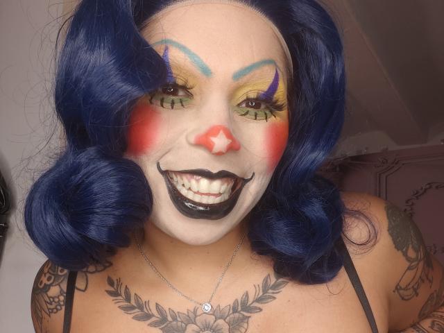Chili with dark blue hair and clown makeup, black lipstick, and yellow eyeshadow, grinning widely, showing white-painted face and tattoos.