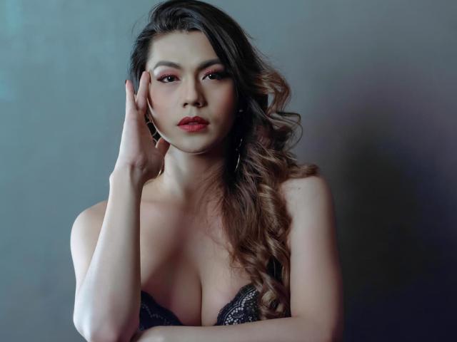 ShelandWilliams with wavy brown hair, pink eyeshadow, and red lipstick, touching her face, wearing a black lace bra, showing her big tits, against a gray background.