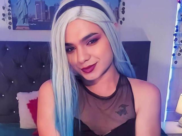 IsabellaDark with white-blue hair, black mesh top, red lipstick, and tattoo, smiling at camera. Boobs visible under top. Bedroom with dark headboard and decorative lights.