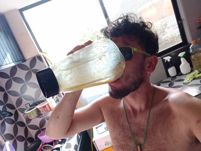 DavydWolfy69, com cabelo castanho encaracolado, barba e colar verde, bebendo de um recipiente grande, espumoso e amarelo com tampa preta, sem camisa, em uma cozinha..