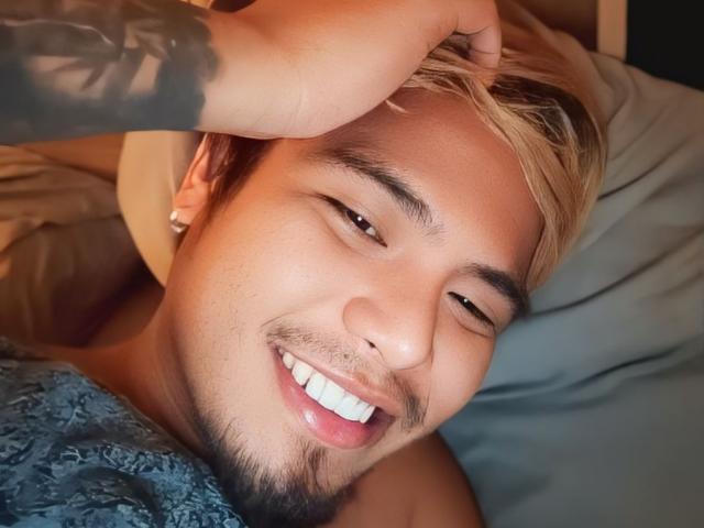 UrAsianMister with blonde hair, light brown skin, and a cute smile, winking and holding his head with a tattooed arm, lying on a bed with a blue patterned shirt.