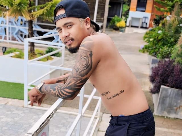 Shirtless UrAsianMister with tattoos, black cap, and dark pants, leaning on a railing, showing a tiger tattoo on his right arm and a small tattoo under his left rib.