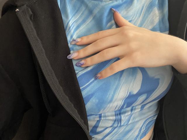 La mano di Valissia, con le unghie decorate di glitter viola e bianchi, riposa sulla sua maglietta blu tie-dye, che mette in mostra i suoi piccoli seni. Indossa una giacca nera parzialmente aperta. La sua pelle è chiara..
