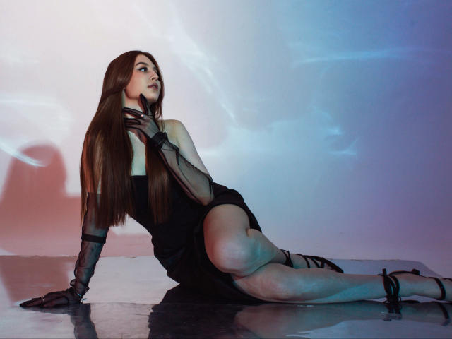 EllyCat with long brown hair, black dress, sheer gloves, and strappy heels, sitting with one leg up, hand on neck, looking up, showing her ass and legs.