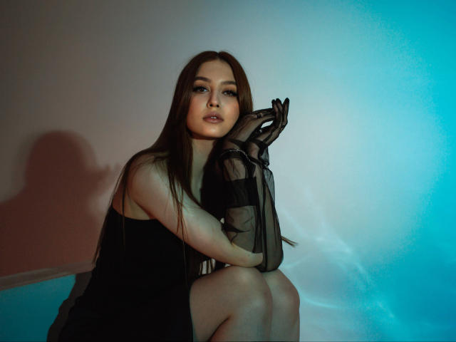 EllyCat with long brown hair, wearing a black sheer dress and gloves, sits with knees up, looking at camera, blue and pink light on right, shadow on left.