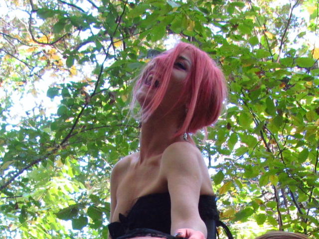 MissCelestine with pink bob, looking up, exposing her bare shoulders and black strap dress, green leaves in background, sun filtering through.