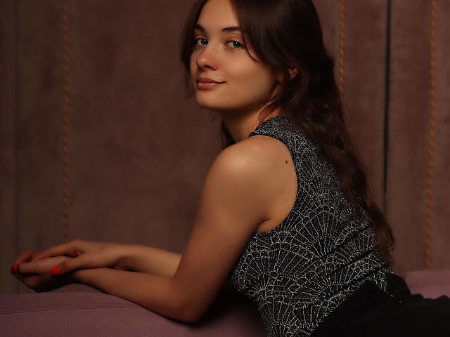 LuiMartin with long brown hair, wearing a sleeveless black and white patterned top, smiling slightly, leaning on a pink surface, red nail polish, cute face, soft light.