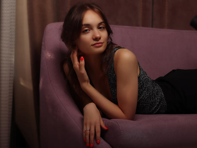 LuiMartin with red nails, lying on a purple couch, wearing a black patterned top, looking at the camera with a slight smile, brown wavy hair, fair skin.