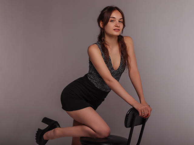 LuiMartin with puffy tits, wearing a deep V-neck, black shorts, and black heels, kneeling on a chair, hands on top, looking at the camera with a slight smile.