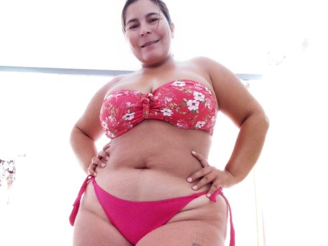 LouiseSexBigBoobs stands in a red floral strapless bikini and matching red panties, showing her curvy, chubby body with large tits and a soft belly, smiling confidently.