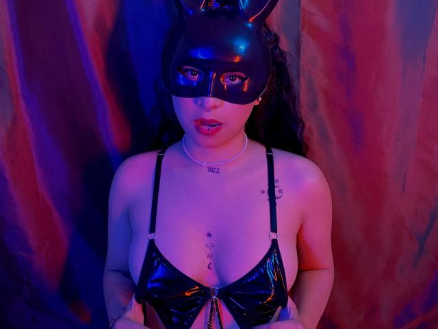 SophieDreamLove draagt een zwart latex masker met konijnenoren, een zwarte glanzende beha en een ketting, met blauwe en roze neonverlichting, waarbij haar borsten en een tatoeage zichtbaar zijn..