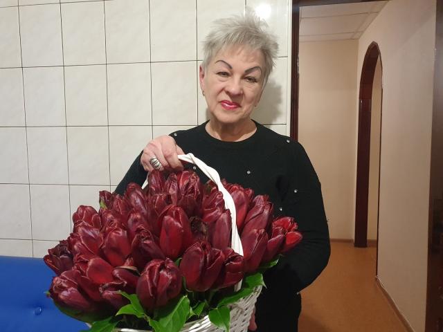 KayleSmoll with short gray hair, black long-sleeve shirt, holding a white basket full of dark red, shiny tulips, smiling, standing in a hallway, wearing a ring.