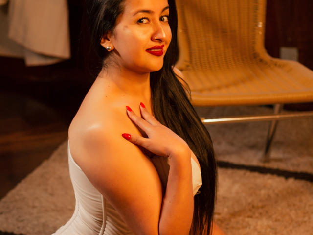 CherryHott69 with long black hair, red lipstick, and red nails, wearing a white strapless top, touching her shoulder, looking at the camera, sitting in a warmly lit room.