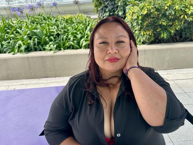 AliceShae, a plus-sized woman with dark red hair, black shirt, and visible cleavage, sits on a purple mat, hand on cheek, red lipstick, and a purple wristband.