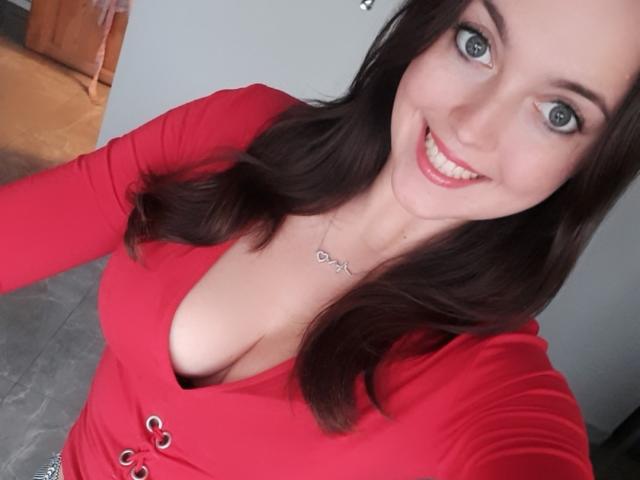 Vanilia69 with blue eyes, smiling, wearing a red V-neck top, showing cleavage, has long brown hair, and a silver necklace with a heart-shaped pendant. Boobs visible.