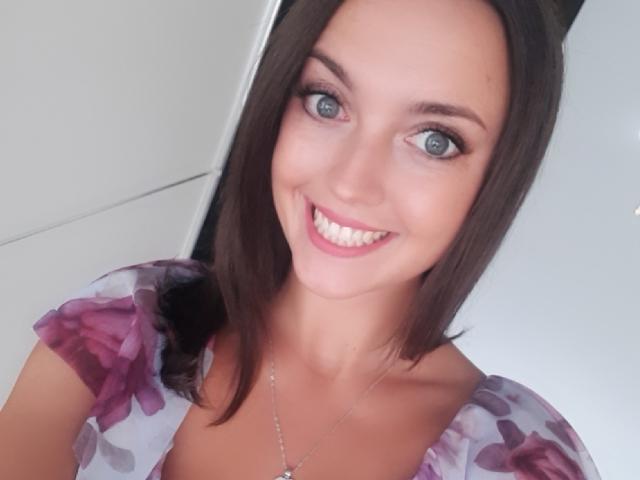 Vanilia69 has blue eyes, a cute smile, and brown shoulder-length hair. She's wearing a purple floral top and a silver necklace. Her tits are small and her ass is not visible.