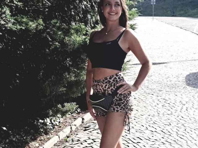 Vanilia69 stands on a cobblestone path, wearing a black sports bra, leopard print shorts, and a patterned clutch, smiling with her hand on her hip, showing her ass.