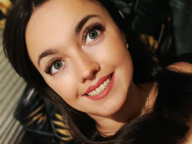 Vanilia69 has green eyes, long black hair, and is smiling with red lipstick. She's wearing a necklace and has subtle makeup with long eyelashes. Her ass is not visible.