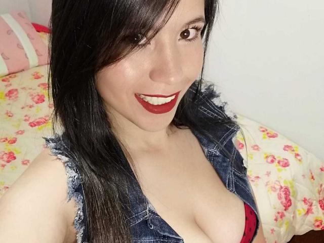LizBellaa, con cabello negro, lápiz labial rojo y una sonrisa, lleva un chaleco vaquero que revela su sujetador rojo y sus grandes senos. Está en una cama con sábanas estampadas con flores..