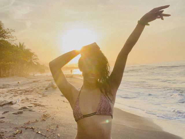 ElizabethMacy in a pink patterned bikini, sun flaring in her eyes, arms raised, wet hair, beach at sunset, water and sand, palm trees in the background, golden light.