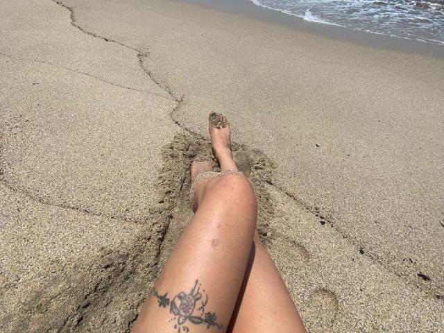 ElizabethMacy's sandy-covered legs with a tattoo on her right thigh, lying on a beach with her feet in the sand and small waves in the background.