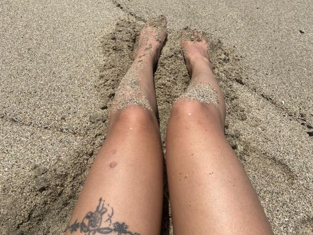 ElizabethMacy's sand-covered legs with a small bruise on the left thigh and a tattoo on the right calf, standing on a sandy beach.