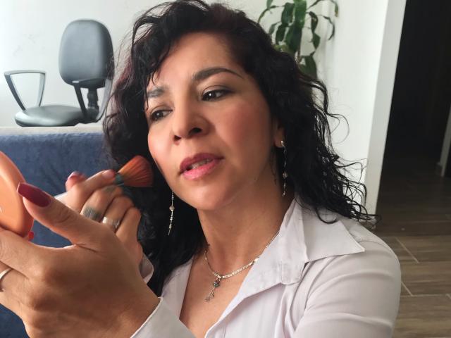 EtoileLatin with curly black hair, medium skin, wearing a white shirt, applying pink lipstick with a brush in her right hand, showing her red-painted nails.