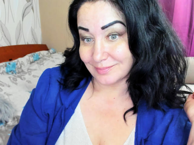 LexyRose has black wavy hair, blue eyes, and is smiling with pink lips. She's wearing a blue shirt over a white top, showing her tits slightly.