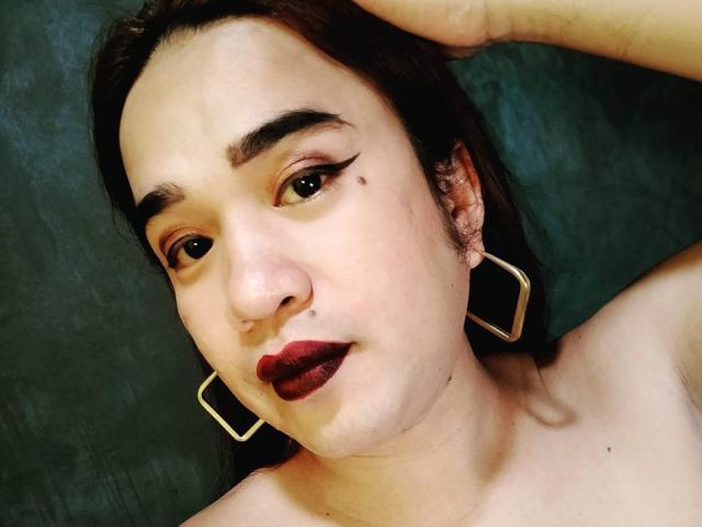 DirtyBigNipples has pale skin, dark eyebrows, red lipstick, black eyeliner wings, a small mole on her right cheek, and wears square gold earrings with her hand in her brown hair.