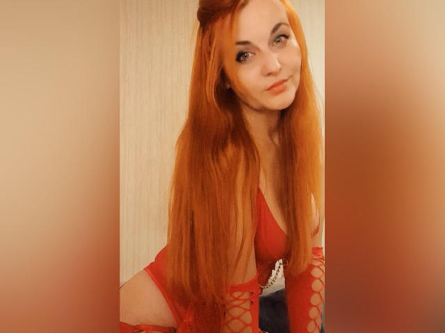 JasminsLove, met lang rood haar, draagt rode lingerie met veters en heeft een schattige glimlach. Ze leunt voorover, waarbij haar rode bh en bijpassende armen zichtbaar zijn, met een sexy uitstraling..
