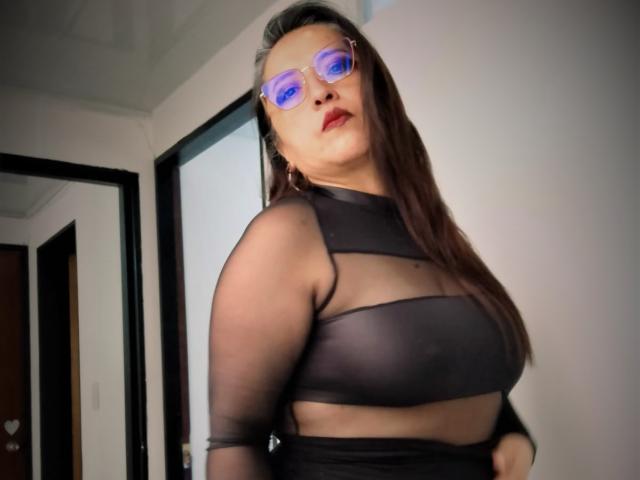 PaolaLady with blue reflective glasses, wearing a sheer black top, exposing her big tits and showing her nipples, has long brown hair and red lipstick.